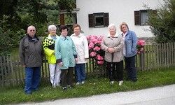 Eine Gruppe älterer Menschen in passenden Outfits steht vor einem Haus mit rosa Blumen und einem Holzzaun.