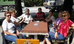 Eine Gruppe von Menschen sitzt an einem Picknicktisch, einige tragen Sonnenbrillen. Eine braune Tischdecke liegt auf dem Tisch, und ein Lkw ist im Hintergrund geparkt.