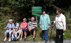 Eine Gruppe älterer Menschen sitzt auf einer Bank mit einem grünen Schild dahinter. Zwei Personen stehen in der Nähe, eine hält einen Stock.