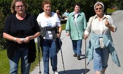 Vier Frauen, alle in Jeans gekleidet, gehen auf einem gepflasterten Weg. Eine Frau benutzt einen Stock und eine andere hat ein Tuch um den Hals. Im Hintergrund befinden sich Bäume und eine grasbewachsene Fläche.