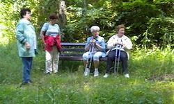 Vier Frauen sind draußen. Zwei sitzen auf einer Bank, eine steht mit einer roten Jacke und die vierte steht in der Nähe. Sie sind in einem Grasbereich mit Bäumen im Hintergrund.
