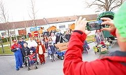 Eine Person fotografiert eine Parade von Menschen in Kostümen, darunter Kinder in Tierkostümen, die mit Kinderwagen und Karren die Straße entlanggehen.