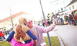 Eine Person mit rosa Perücke und Brille macht ein Selfie mit einem pelzigen Plüschtier vor einer belebten Straße.