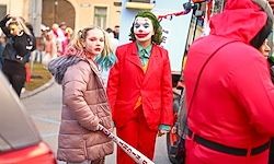 Zwei junge Frauen in Joker-Kostümen halten einen Baseballschläger in einer Straße mit einer Menschenmenge und geparktem Auto.