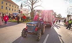 Eine Parade mit einem blauen Traktor und einem rosa Anhänger, auf dem zwei Personen sitzen. Zuschauer säumen die Straße, mit Gebäuden und Bäumen im Hintergrund.