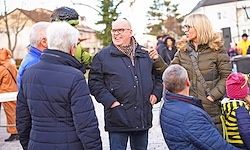 Eine Gruppe von Menschen, darunter zwei Erwachsene, die sich unterhalten, steht draußen in einer kühlen Umgebung. Sie sind in warme Mäntel gehüllt und scheinen in ein freundliches Gespräch vertieft zu sein.