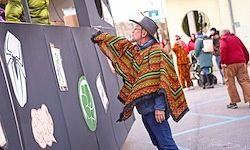 Ein älterer Mann in einem farbenfrohen Poncho zeigt auf eine Wand mit verschiedenen Aufklebern. Im Hintergrund gehen Menschen an einem Gebäude vorbei.