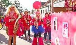 Eine Gruppe von Menschen in roten und pinkfarbenen Kostümen geht die Straße entlang, spielt Musikinstrumente, einer hält einen roten Luftballon.
