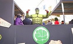 Eine Person in einem Hulk-Kostüm steht auf einer erhöhten Plattform, umgeben von einer Menge Zuschauer, mit einem Gebäude und einem grünen Faustlogo im Hintergrund.
