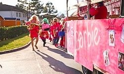 Eine Gruppe von Menschen in rosa und grünen Kostümen marschiert auf einer Straße, mit einem rosafarbenen Wagen dahinter. Auf dem Wagen steht das Wort Barbie.