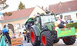Ein Traktor, geschmückt mit Luftballons und einem Schild mit der Aufschrift 'Royal Lassing', fährt die Straße hinunter. Eine Person mit blauen Haaren hält einen Korb. Gebäude säumen die Straße.