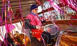 Ein Mann in einem roten Jackett spielt eine Trommel auf einem Boot mit rosa Streamern. Eine weitere Person sitzt in der Nähe, und Menschen sind im Hintergrund zu sehen.