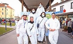 Eine Gruppe von Personen in weißen Schutzanzügen steht vor einem Gebäude. Die Männer sind auf der rechten Seite, die Frauen auf der linken. Sie tragen alle Masken. Dahinter laufen Menschen auf der Straße.