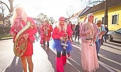 Eine Gruppe von Menschen in pinkfarbenen Kostümen geht eine Straße entlang und spielt Instrumente wie Tuba und Trompete. Gebäude und Autos sind auf beiden Seiten der Straße zu sehen.