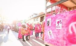 Eine Gruppe von Menschen in rosa Kostümen und Perücken geht auf einer Straße, mit einem rosa Wagen und Bildern dahinter.