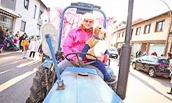 Eine Person in einer rosa Jacke fährt einen blauen Traktor mit einem Hund auf dem Schoß. Im Hintergrund sind eine Menge Leute und Autos zu sehen.