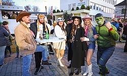Eine Gruppe von Menschen in verschiedenen Kostümen, darunter ein Cowboy, eine Hexe und ein Superheld, posiert für ein Foto auf einer Kopfsteinpflasterstraße.
