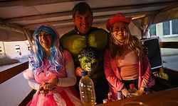 Drei Personen in Kostümen posieren für ein Foto in einem Fahrzeug. Ein Mann in der Mitte mit einem grün-schwarzen Kostüm und zwei Frauen an den Seiten, eine mit blauen Haaren.