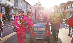 Eine Gruppe von Menschen in Kostümen, einschließlich eines Mannes in Pink mit einer Trompete, reitet auf einem blauen Traktor bei einem Straßenumzug.