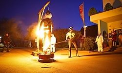 Ein Mann in einem Kostüm schwingt eine Fackel über ein Lagerfeuer, während andere nachts vor einem Gebäude stehen.