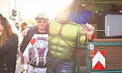 Ein Mann in einer Hulk-Kostüm posiert für ein Foto mit einem Mann in einem schwarzen Shirt, Hut und Sonnenbrille. Sie stehen neben einem grünen Wagen mit einem Lautsprecher und einem roten und weißen Dreiecksschild.