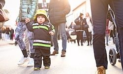 Ein kleines Kind in einem Feuerwehrmann-Outfit läuft mit einem Elternteil auf dem Bürgersteig, umgeben von anderen Fußgängern mit Kinderwagen.