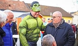 Ein Mann in einem grünen Kostüm interagiert mit einem älteren Mann. Dahinter versammelt sich eine Menschenmenge um Gebäude. Eine Person trägt eine Brille.