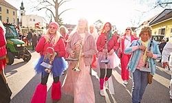Eine Gruppe von Frauen in pinken und weißen Kostümen, einschließlich Stiefeln, geht mit Trompeten und Handtaschen die Straße entlang. Sie nehmen wahrscheinlich an einem Umzug teil.