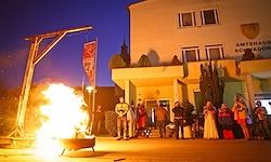 Ein brennendes Feuer lodert vor einem Gebäude, mit einem Galgen in der Nähe. Menschen in Kostümen versammeln sich um das lodernde Feuer, das bei Nacht unter einem Sternenhimmel leuchtet.