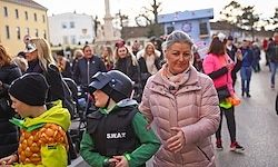 Eine Frau und zwei Kinder gehen während einer Veranstaltung im Freien mit anderen Menschen die Straße entlang. Sie sind von Gebäuden und anderen Teilnehmern umgeben.