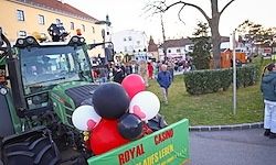 Ein dekorierter Traktor mit roten, weißen und schwarzen Ballons und einem Schild 'Royal Casino' steht auf der Straße, mit Menschen und Gebäuden im Hintergrund.