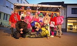 Eine Gruppe von Menschen in bunten Kostümen, einschließlich Superhelden, posiert für ein Foto vor einem Gebäude mit einer überdachten Struktur.