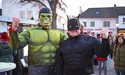 Zwei Personen, die als Superhelden verkleidet sind, einer als Hulk und der andere als Batman, stehen gemeinsam auf einer Straße mit Gebäuden und Menschen im Hintergrund.