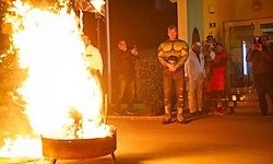 Ein Mann steht bei einer großen Feuerstelle in der Nacht mit anderen um ihn herum, einige machen Fotos.