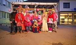 Eine Gruppe von Menschen in pinken Kostümen und Masken posiert für ein Foto vor einem Gebäude bei Nacht.