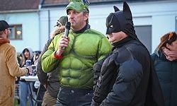 Zwei Männer in Kostümen, einer als Hulk und der andere als Batman, stehen vor einer Menge. Der Hulk hält ein Mikrofon und spricht, während Batman neben ihm steht.