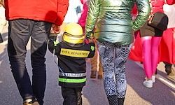 Ein Kleinkind, das als Feuerwehrmann gekleidet ist, geht mit zwei Erwachsenen, einem in einem roten Mantel und dem anderen in einem grünen Puffer. Sie sind in einer Parade mit Menschen, die bunte Strumpfhosen tragen.