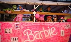 Eine Gruppe von Menschen in Kostümen posiert für ein Foto in einem Barbie-Thema. Sie stehen auf einer rosa Wand mit verschiedenen Bildern und dem Wort Barbie.