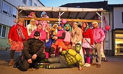 Eine Gruppe von Menschen in verschiedenen farbenfrohen Kostümen, darunter eines in einem grünen Superheldenkostüm, posiert für ein Foto vor einem Gebäude.