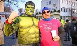 Zwei Männer stehen auf einer Straße, einer als Hulk verkleidet und mit einem Becher in der Hand, der andere in einem rosa T-Shirt. Sie sind von Menschen umgeben. Gebäude mit Pflanzen sind im Hintergrund.