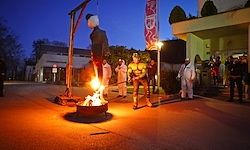 Eine Menschenmenge versammelt sich um ein Lagerfeuer, mit einer Person, die von einem Galgen hängt und einem Feuer darunter. Die Szene wird vom Licht des Lagerfeuers beleuchtet, das Schatten wirft.