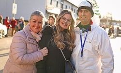 Drei Personen posieren für ein Foto im Freien, wobei eine Person einen weißen Schutzanzug und einen Helm trägt. Die Frau mit Brille steht zwischen zwei Frauen, von denen eine lächelt.