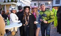 Fünf Menschen in Kostümen posieren für ein Foto auf einer Stadtstraße. Die Frau in der Mitte trägt einen langen schwarzen Mantel. Der Mann rechts hält ein Getränk in einem Glas.