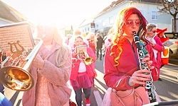 Eine Straßenparade mit Musikern in pinken Kostümen, darunter eine Frau, die eine Klarinette und eine andere, die eine Trompete spielt. Im Hintergrund scheint die Sonne hell.