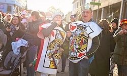 Menschen versammeln sich auf einer Straße für eine Veranstaltung, zwei Männer in Spielkarten-Thema-Kleidung interagieren mit der Menge, einer hält ein Getränk.