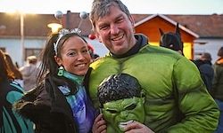 Ein lächelndes Paar auf einer Kostümparty, der Mann in einem Hulk-Kostüm und die Frau mit einer Krone. Sie sind von anderen Menschen umgeben.