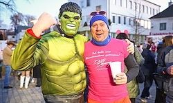 Zwei Männer in Kostümen, einer als Hulk und der andere in Pink, stehen zusammen auf einer belebten Straße, halten einen Becher und posieren für ein Foto.