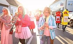 Eine Gruppe von Frauen in rosa Tüllröcken und Brillen spielt Musikinstrumente in einer Parade. Sie sind von anderen Teilnehmern umgeben, einer trägt ein gelbes Maskottchenkostüm. Gebäude säumen die Straße hinter ihnen.