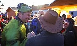 Zwei Männer, einer mit grüner Maske und der andere mit Cowboyhut, lachen und schütteln sich die Hände in einer Menschenmenge. Dahinter schaut eine Frau mit Bandana und Brille zu.