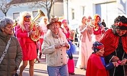 Eine Gruppe von Menschen in roten Kostümen, einige mit Hüten, geht auf der Straße. Eine Frau vorne hält eine Tuba, und andere spielen verschiedene Musikinstrumente. Die Straße ist von Gebäuden gesäumt.